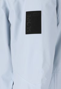 Ljusblå vattentät jacka med en svart rektangulär logotyp på ärmen. Slät textur, minimalistisk design och inga synliga knäppningar.