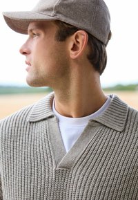 Lysegrå strikket sweater med ribbet krave og v-hals, lagdelt over en hvid t-shirt. Personen bærer en lysegrå kasket.