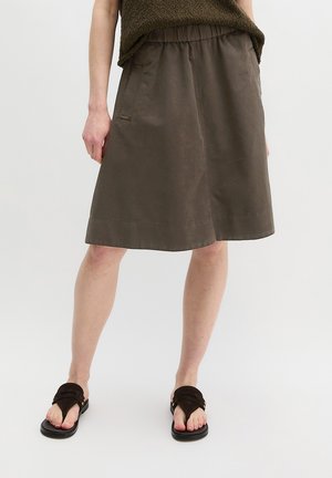 Brązowa spódnica do kolan z elastycznym paskiem, boczne kieszenie, noszona z ciemnobrązowymi sandałami na jasnym tle.