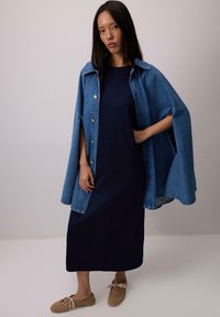 Ženska, oblečena v midi obleko temno modre barve z modrim jeans plaščem in bež balerinkami, ki stoji pred enolično svetlo sivo ozadje.