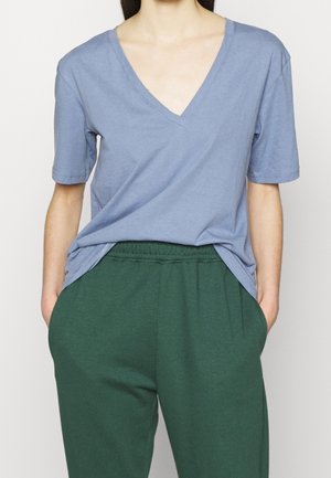 Personne portant une chemise bleue ample à col en V et manches courtes, rentrée dans un pantalon vert à taille élastique, les mains dans les poches.