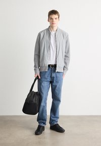 Jachetă bomber gri cu mâneci în contrast, tricou alb, blugi blue, adidași negri, ținând o geantă din țesătură neagră pe un fond deschis.