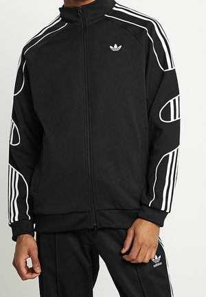 Homme portant une veste de survêtement Adidas noire avec des rayures blanches et des logos, assortie à un pantalon de survêtement Adidas noir.