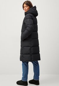 Zwarte pufferjas met een capuchon, voorzien van een geweven ontwerp, zijzakken en een rond logo-patch op de bovenarm. Gepaarde met blauwe jeans en laarzen.