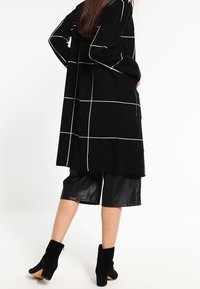 Svart lång kappa med vit rutmönstring, kombinerad med glänsande svarta lädershorts och ankelskor. Mjuk textur med avslappnad passform.