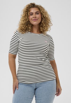 Kobieta o kręconych blond włosach, ubrana w czarno-białą koszulkę w paski z krótkim rękawem i jasnoniebieskie dżinsy, uśmiechająca się do aparatu.