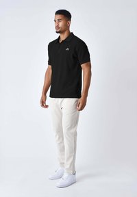 Camisa polo negra de canalé con mangas cortas y un pequeño logo bordado, combinada con pantalones beige claro y zapatillas blancas.