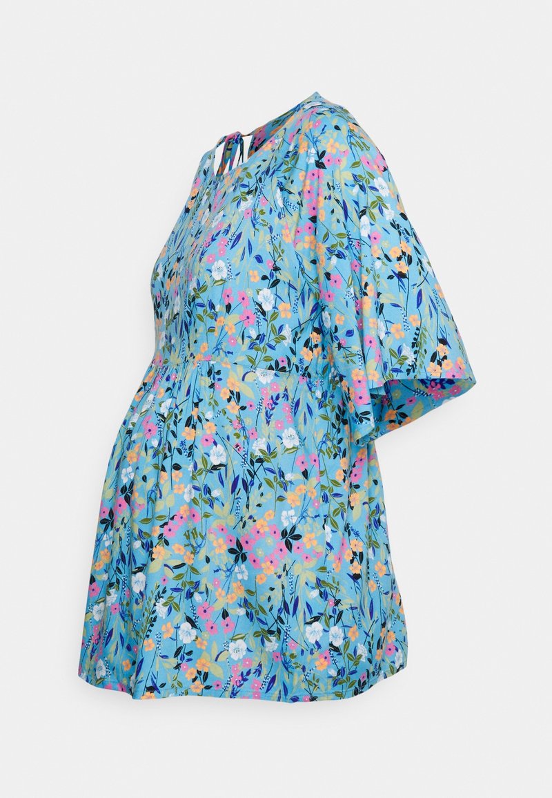 Blouse de maternité bleue à fleurs avec manches courtes et détail noué au niveau de l'encolure dos sur fond uni.