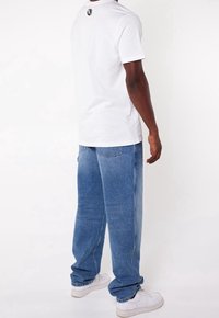 Vit bomullströja med rund hals, kombinerad med ljusblå denimjeans. Noterbar logotypdetalj på baksidan av tröjan, vita sneakers.