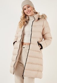 Béžová péřová bunda se zipem vpředu, s kapucí vlněnou z kožešiny, horizontálním prošíváním a postranními kapsami, kombinována s lehkým svetr.