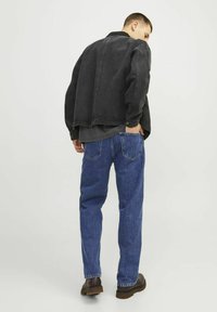 Chaqueta de mezclilla negra con un corte holgado, colocada sobre una camisa oscura, combinada con jeans rectos azules y botas marrones.