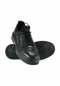 Czarne skórzane sneakersy na sznurówki z teksturowanymi gumowymi podeszwami, perforowanym przodem i dzianinowymi kołnierzami wokół kostki, ustawione tak, że jeden but stoi, a drugi spód jest skierowany do góry.