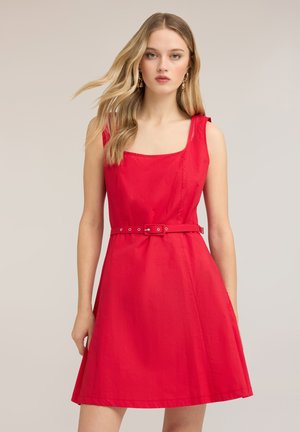 Junge Frau mit langen blonden Haaren, die ein ärmelloses rotes Kleid mit quadratischem Ausschnitt und einem dünnen passenden Gürtel in der Taille trägt.