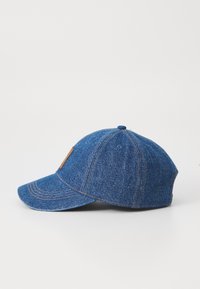 Denim caps med buet skjerm, en blå teksturert overflate, og detaljer i brunt lær. Synlige sømmer langs sømmene.