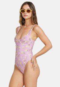 Maillot de bain une pièce en violet clair avec un motif floral, texture côtelée, bretelles larges et design à dos échancré, associé à des lunettes de soleil jaunes.
