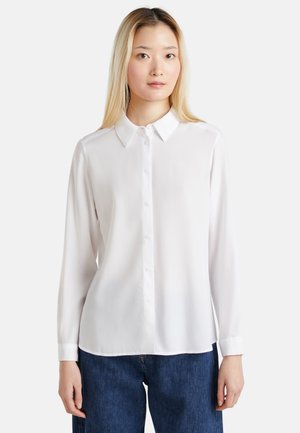 Junge Frau mit glatten blonden Haaren, die ein weißes Hemd mit Knöpfen und blaue Jeans trägt, steht vor einem einfachen hellen Hintergrund.