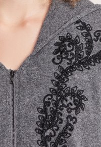 Sudadera gris con cremallera que presenta un intrincado bordado negro de hojas y enredaderas en un lado de la cremallera. Tejido suave y textura agradable.