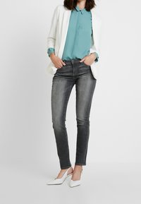 Femme portant un blazer blanc, un chemisier turquoise, un jean skinny noir délavé et des escarpins blancs à bride arrière pointus, debout les mains dans les poches.