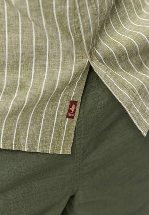 Close-up van de zoom van een groen gestreept overhemd over effen olijfgroene broek, met een klein rood stoffen label met een gouden logo.