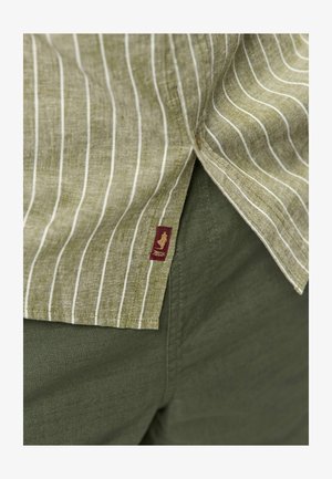 Roheline triibuline särk, mille all paistab ühevärvilised oliivrohelised püksid, millel on väike punane kangast silt kuldse logoga.