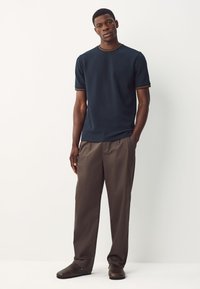 Camicia marina a maniche corte con sottile rifinitura marrone a contrasto, abbinata a pantaloni beige a gamba dritta e scarpe marrone scuro, con linee semplici.