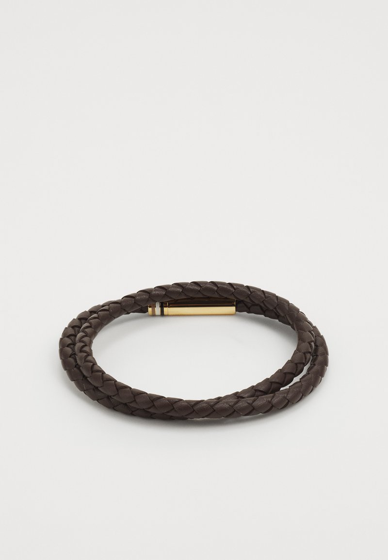 Apsiūtas tamsiai rudos odos apyrankė su auksinės spalvos užsegimu, turinčiu horizontalių juostelių. Lygus paviršius ir lanksti konstrukcija.