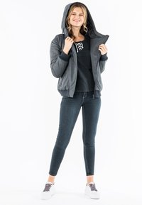 Graue Bomberjacke mit Kapuze, gestrickten Bündchen und Reißverschlussdetails, getragen über einem schwarzen Shirt und kombiniert mit dunklen Jeans und weißen Turnschuhen.