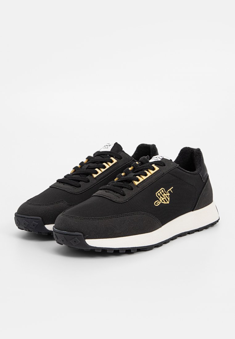 Svarta sportiga sneakers med ett strukturerat tygöverdrag, vit mellansula och gyllene detaljer. Har snörning och en gummisula för grepp.
