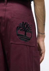 Gros plan sur la poche d'un pantalon bordeaux avec un logo d'arbre brodé en noir, montrant une partie d'un bras tatoué à côté.