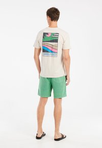 Beige T-shirt med farverig "Zurriola Beach" grafik på ryggen; grønne shorts; person iført sorte flip-flops, stående på en ensfarvet baggrund.