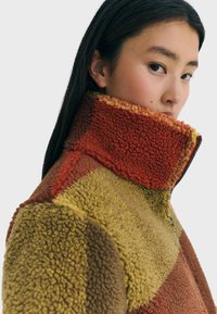 Una joven lleva una chaqueta de forro polar de cuello alto y multicolor en tonos de rojo, amarillo y marrón, mirando de lado con una expresión neutral.