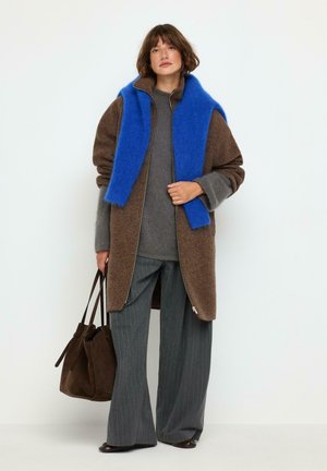 Femme portant un manteau marron par-dessus un pull gris et un pantalon à fines rayures, une écharpe bleu vif drapée sur les épaules, tenant un sac à main marron foncé.