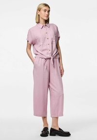 Mono de lino rosa con mangas cortas, botonera al frente, dos bolsillos y cintura anudada. Combinado con mocasines negros. Parte delantera plana con piernas anchas.