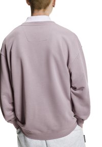 Lila sweatshirt met een relaxte pasvorm, ronde hals, oversized mouwen en subtiele stikdetail aan de achterkant. Gecombineerd met grijze joggingbroek.