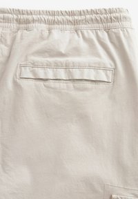 Pantaloni corti color beige chiaro in cotone con vita elasticizzata e una tasca posteriore con pattina. Texture morbida e design semplice.