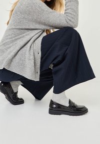 Femme accroupie, portant un pull gris, un pantalon large bleu marine, des chaussettes grises et des mocassins noirs brillants sur un fond clair uni.