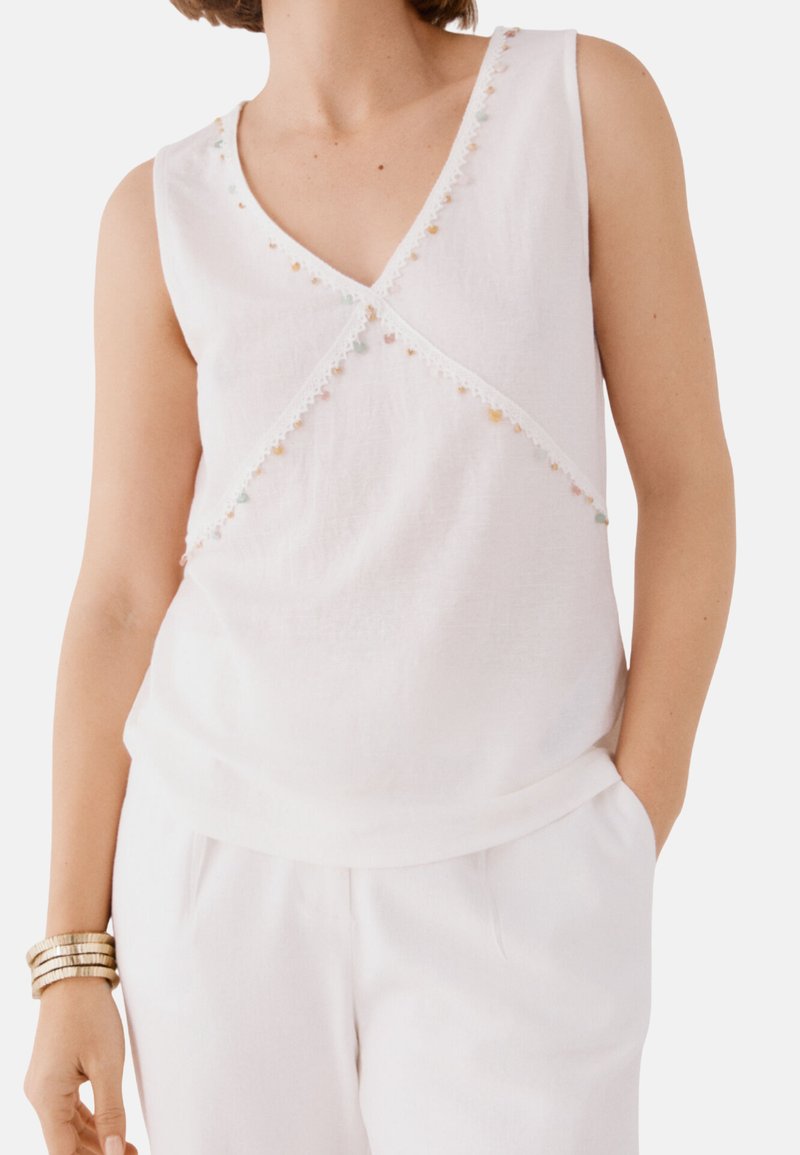 Witte mouwloze top met een V-hals, voorzien van geborduurde accenten langs de halslijn en een textuurstof. In combinatie met witte broeken.