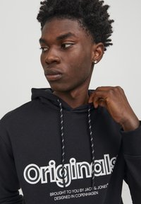 Sudadera negra con la palabra "Originales" en letras blancas en negrita, con tejido texturizado y cordones en contraste. Diseño sencillo, ajuste casual.