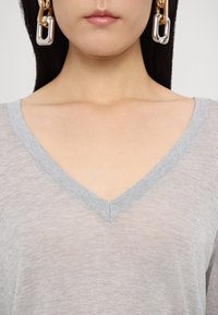 Haut gris à col en V avec une texture douce, doté d'un ourlet fin et d'une coupe décontractée. Un mannequin arbore de grandes boucles d'oreilles en chaîne métallique.