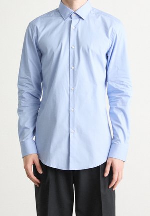 Mężczyzna ubrany w jasnoniebieską, długorękawową koszulę na guziki oraz czarne spodnie, stojący na tle jednolitego białego tła.