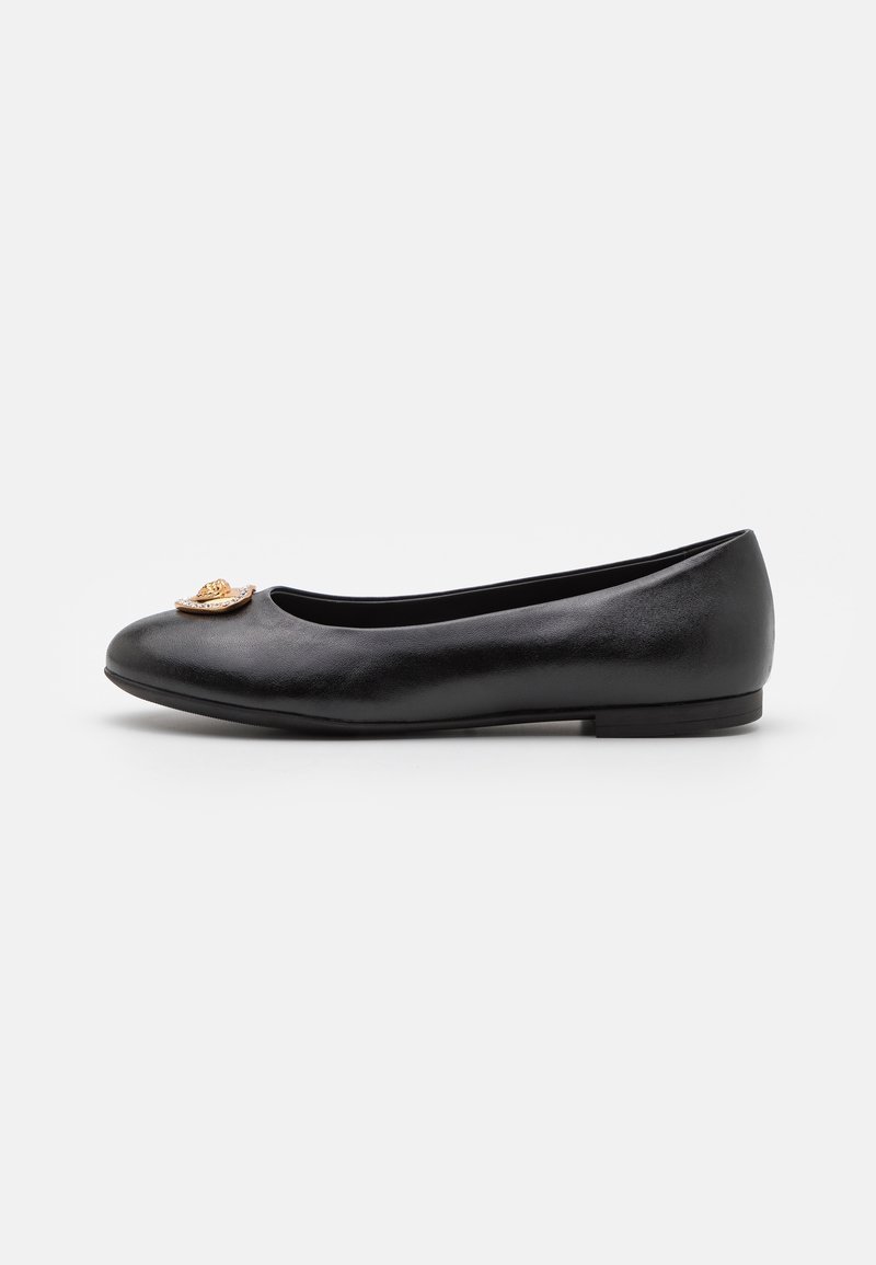 Chaussure plate en cuir noir avec bout rond et emblème décoratif doré à l'avant, vue de côté sur fond blanc.