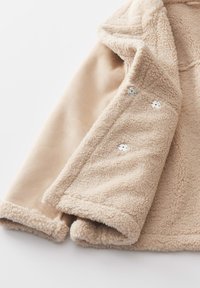 Veste en polaire beige avec un extérieur texturé et duveteux et des manches lisses, ornée de boutons-pression argentés sur la fermeture avant.