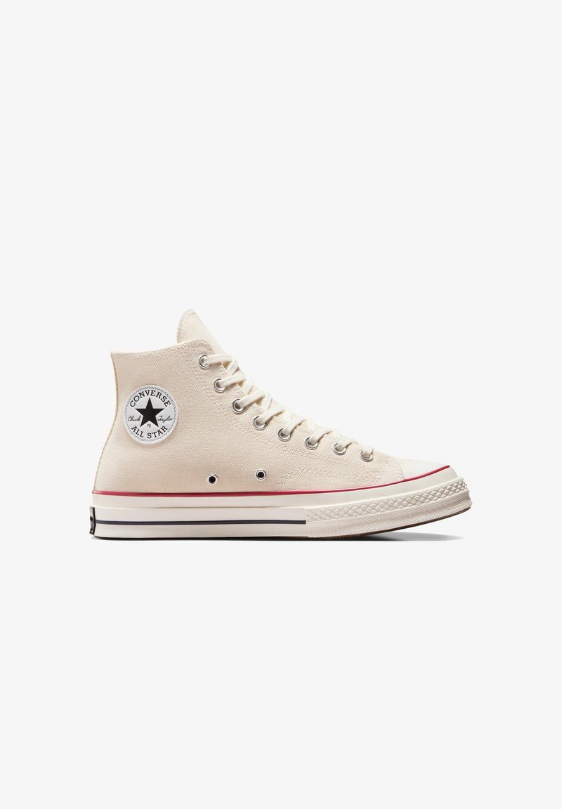 Baskets montantes en toile beige avec un embout en caoutchouc, un écusson logo noir, des œillets en métal et une bande rouge d'accent sur la semelle.