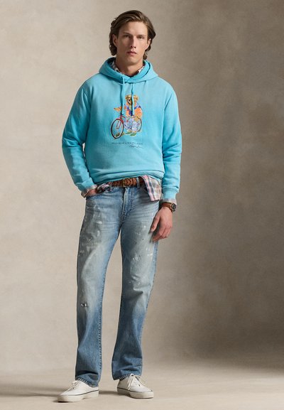 Sweat à capuche turquoise avec un motif d'ours, jean bleu clair délavé, baskets blanches. Le sweat à capuche est doté d'un cordon de serrage et de poignets côtelés.