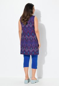 Ärmellose lila Tunika mit einem Diamantmuster in Blaugrün und Rot, kombiniert mit blauen Crop-Leggings und hellblauen Slippern.