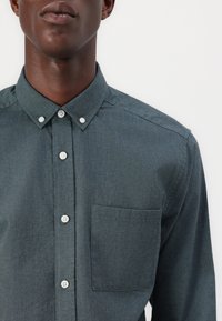 Button-up skjorte i mørk teal stof med en glat tekstur. Har en spids krave, hvide knapper og en brystlomme.