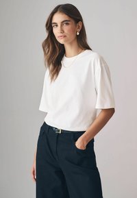 Weißes Baumwoll-T-Shirt mit kurzen Ärmeln, kombiniert mit hoch sitzenden schwarzen Hosen. Eine dünne Goldkette und schlichte Ohrringe runden das Outfit ab.