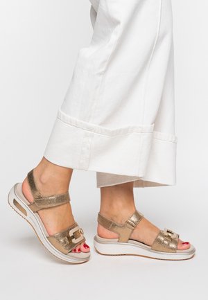 Feet wearing metallic gold sandals with white soles and adjustable straps, paired with wide-legged white pants.