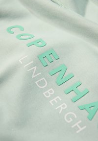 Tejido verde menta con letras en relieve "COPENHAG" en aqua y "LINDBERGH" en blanco. Textura suave con un ligero brillo.
