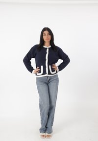 Cárdigan marino con ribete blanco y cierres de botones, que cuenta con dos bolsillos frontales. Combinado con jeans de tiro alto en azul claro y sandalias.